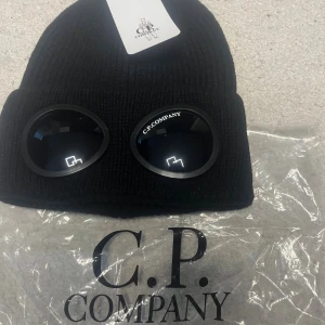 Svart C.P. Company mössa med goggles - Svart ribbstickad mössa från C.P. Company med ikoniska goggles framtill. Snygg och unik design som sticker ut, perfekt för dig som gillar streetwear och vill ha något extra. Tillverkad i mjukt stickat material som håller dig varm. Kvitto finns