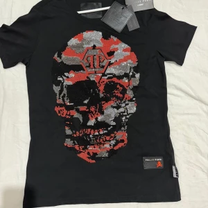 Philip Plein T-shirt  - Märke: Philipp Plein Modell: Homme kollektion Färg: Svart med flerfärgat stentryck Patch med Philipp Plein-logga nertill på framsidan Märkestags hänger fortfarande kvar Äkthetsband med logga i sidosömmen