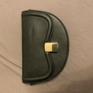 Svart kuvertväska med gulddetalj - Stilren svart kuvertväska i skinnimitation med snygg guldfärgad låsdetalj framtill. Väskan har en halvmåneformad design och ljus insida. Perfekt för att bära det viktigaste när du vill hålla det enkelt och classy.