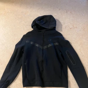 Nike tech fleece  - Svart Nike tech fleece storlek 158-170 så xs. Jackan har hel dragkedja, huva och en diskret ficka med dragkedja på ena ärmen. Snygga svarta detaljer över bröstet och en sportig look. Perfekt för dig som gillar streetwear och sportig stil. Pris går att diskutera vid snabb affär