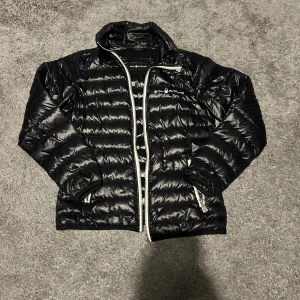 Svart pufferjacka från Sail Racing - Bra skick förutom att dragkedjan har problem och funkar inte så bra men säkert lätt att fixa Svart pufferjacka från Sail Racing med blank finish och horisontella quiltade sömmar. Jackan har dragkedja framtill, hög krage och logga på bröstet. Perfekt för kyliga dagar och har en sportig vibe.