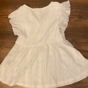 Vit kortklänning med volangärmar - Supersöt vit kortklänning med broderade detaljer och volangärmar. Klänningen har v-ringning med spetskant och ett lätt, luftigt tyg som känns perfekt för varma dagar. Den har en lös passform och ett romantiskt uttryck.