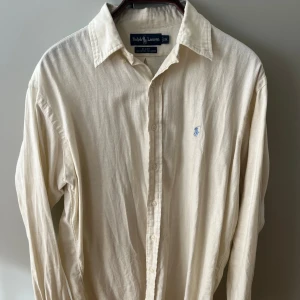 Beige linneskjorta från Ralph Lauren - Klassisk beige skjorta från Ralph Lauren i mix av bomull och linne. Skjortan har lång ärm, knappar framtill och ikonisk broderad logga på bröstet. Perfekt oversized fit för en chill och stilren look. Passar mer som L då den är lite oversized