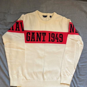Vit och röd GANT tröja med tryck - Vit stickad tröja från GANT med bred röd rand över bröstet och armarna, samt svart text 'GANT 1949' framtill. Rund halsringning och ribbade muddar. Snygg och sportig collegekänsla, perfekt för dig som gillar klassisk stil.