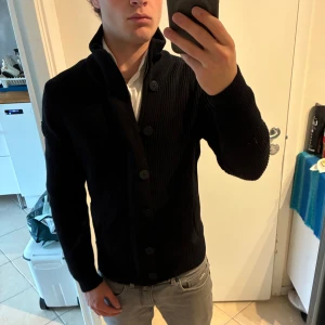 Svart kofta från Marina Yachting - Snygg svart ribbad kofta från Marina Yachting med hög krage och stora knappar framtill. Tröjan är stickad i ull och har långärmad passform. Perfekt för dig som gillar stilrena och klassiska plagg med lite edge. Bra skick 