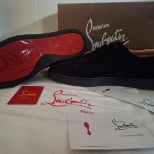 Christian Louboutin svarta sneakers - Svarta sneakers från Christian Louboutin i mocka med klassisk röd sula och rund tå. Skorna har svarta snören och diskreta detaljer, samt beige innersida. Perfekta för dig som vill ha en lyxig och stilren look med ikonisk touch.
