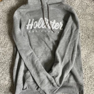 Grå hoodie från Hollister - Säljer en grå hoodie från Hollister med vit broderad logga på bröstet. Tröjan har huva med snörning, magficka och långa ärmar. Perfekt för chill dagar eller när du vill vara extra bekväm.