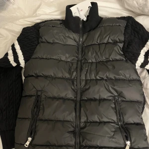 Svart pufferjacka från Moncler - Svart pufferjacka från Moncler med stickade ärmar och vita ränder på armarna. Jackan har hög krage, två fickor med dragkedja och quiltad front. Materialmix av polyester och stickat ger en cool kontrast. Perfekt för dig som gillar streetstyle och vill ha något unikt.