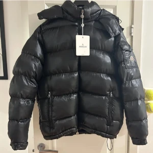 Svart Moncler maja - Säljer en fet svart pufferjacka från Moncler med glansig finish och klassisk logga på ärmen. Jackan har huva, dragkedja framtill och två sidofickor med dragkedja. Perfekt för kalla dagar och har en cool comic patch på insidan scannern funkar pris kan pratas om