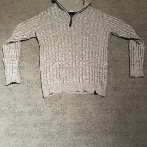 Grå stickad polotröja med half zip - Snygg beige stickad polotröja från Morris med halv dragkedja och högt krage. Tröjan har ett strukturerat mönster på framsidan och ärmarna, samt ribbade muddar vid ärmslut och nederkant. Perfekt för dig som gillar klassisk stil med en modern twist.