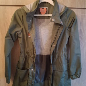 Olivgrön parkajacka med huva - Snygg olivgrön parkajacka från Girl Gang med grått innerfoder i huvan. Jackan har dragkedja och tryckknappar framtill, två stora fickor och resår i midjan för en schysst passform. Perfekt för vår och höst! Size 140