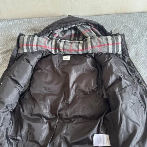 Svart pufferjacka från Burberry - Säljer en svart pufferjacka från Burberry med avtagbar huva och klassiskt rutigt foder i grått, vitt och rött. Jackan har dragkedja, knappar och två sidofickor med zip. Perfekt för kalla dagar med sin quiltade design och snygga logga på ärmen. Man kan göra jackan till en väst också 
