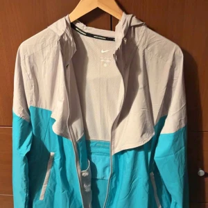 Nike turkos och grå vindjacka - Nike vindjacka i turkos och ljusgrå med snyggt färgblockat mönster. Jackan har huva, hel dragkedja och två sidofickor med vita detaljer. Perfekt för dig som gillar sportig stil och vill sticka ut med fräscha färger.