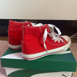 Röda höga sneakers med vit sula - Snygga röda sneakers i klassisk high-top modell med vit gummisula och tå, vita snören och kontrastsömmar. Skorna har rund tå och är tillverkade i canvas. Perfekta för dig som vill sticka ut med färg på fötterna.