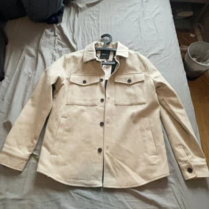 Jack&jones overshirt jacka - Stilren beige overshirt jacka från Jack & Jones med klassisk krage, två bröstfickor med knappar och raka ärmar. Jackan är tillverkad i en ullblandning och har svarta knappar framtill. Perfekt för lager-på-lager och snygg till jeans.