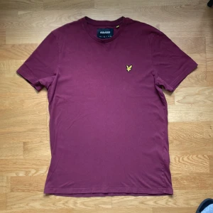 Vinröd t-shirt från Lyle & Scott - Snygg vinröd t-shirt från Lyle & Scott med klassisk rund hals och korta ärmar. På bröstet sitter den ikoniska broderade gula örnen. T-shirten är gjord i mjuk bomull och har en clean, stilren look. 