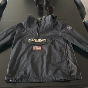Svart vindjacka från Napapijri - Säljer en svart vindjacka från Napapijri med huva och stor ficka framtill. Jackan har dragkedja upptill, norsk flagga och logga på bröstet samt patch på ärmen. Perfekt för blåsiga dagar och snygg streetstil. Pris kan diskuteras 
