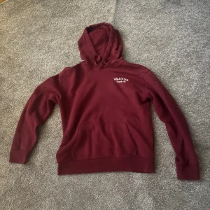 Vinröd hoodie från Hollister - Vinröd hoodie från Hollister med vit logga på bröstet och stor text på ryggen. Klassisk känguruficka framtill, huva med snörning och mjukt material som känns skönt mot huden. Perfekt för chill dagar eller när du vill ha en avslappnad stil.