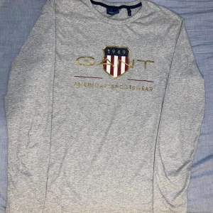 Grå långärmad tröja från Gant - Snygg grå långärmad tröja från Gant med stort tryck framtill i guld, rött och marinblått. Tröjan har rund halsringning med mörkblå kant och klassisk passform. Perfekt för dig som gillar sportig och stilren look.