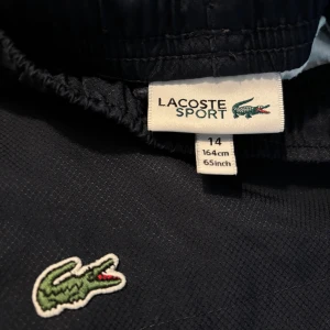 Mörkblå sportshorts från Lacoste - Snygga mörkblå sportshorts från Lacoste med klassisk krokodillogga broderad framtill. Shortsen har elastisk midja och är tillverkade i ett lätt och funktionellt syntetmaterial som passar perfekt för träning eller chill. Enkel design med sidofickor.