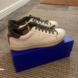 Vita sneakers med gröna detaljer - Snygga vita sneakers med gröna skosnören och mörkgröna detaljer vid hälen. Skorna har perforerat mönster på sidan och klassisk låg profil. Perfekta för en stilren och avslappnad look.