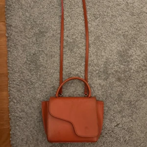 Orange axelväska i skinn med unik form - Snygg orange axelväska i skinn med avtagbar lång rem och ett coolt, asymmetriskt lock. Väskan har även ett praktiskt handtag upptill och detaljer i guldmetall. Perfekt storlek för det viktigaste och ger en trendig touch till din outfit. Defekter finns💓Pris kan diskuteras 💗