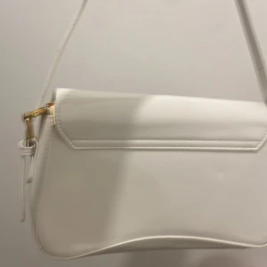 Vit axelväska med gulddetaljer - Snygg vit axelväska med stilren, böjd form och guldfärgade metalldetaljer vid remmarna. Väskan har en slät, glansig yta och minimalistisk design som ger en modern vibe. Perfekt storlek för det viktigaste och enkel att matcha med olika outfits.