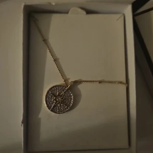 Guldigt halsband Buckley London - Snyggt halsband från Buckley London med ett runt guldigt hänge dekorerat med en stjärna och glittrande stenar. Kedjan är tunn och elegant, perfekt för att lyfta vilken outfit som helst. Kommer i originalförpackning.