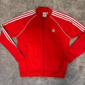 Röd Adidas track jacket med vita ränder - Klassisk röd Adidas track jacket med vita tre ränder längs ärmarna och broderad logga på bröstet. Jackan har hel dragkedja, ribbade muddar och är tillverkad i glansig polyester. Perfekt för en sportig och retro vibe.