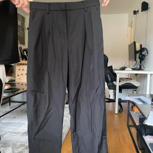 Svarta kostymbyxor med pressveck - Snygga svarta kostymbyxor med klassiska pressveck och hällor för bälte. Byxorna har raka ben och två fickor bak. Perfekta för en clean och stilren look. Materialet känns som bomull eller polyesterblandning.