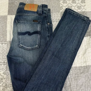 Nudie Jeans blå jeans 27/32 - Snygga blå jeans från Nudie Jeans med klassisk femficksdesign och coola slitningar. Jeansen har en mörk tvätt med ljusare partier och ikonisk broderad bakficka. Modellen är rak och passar perfekt till en avslappnad stil. Knappt använd och har suttit i garderoben 