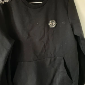 Svart Philipp Plein sweatshirt - Svart sweatshirt från Philipp Plein med silverfärgad logga på bröstet och mönstrad axelband i svart och vitt. Tröjan har rund halsringning och en stor ficka framtill. Tillverkad i mjuk bomull, perfekt för en stilren och lyxig look.