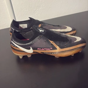 Nike Phantom GX fotbollsskor svart/guld - Säljer ett par Nike Phantom GX fotbollsskor i svart med guldiga och vita detaljer. Skorna har en strumpliknande passform, stora vita swoosh-loggor och texten PHANTOM på sidan. Ovandelen är i syntetmaterial och sulan har fasta dobbar för gräsplan.      Sänker priset vid snabb affär!