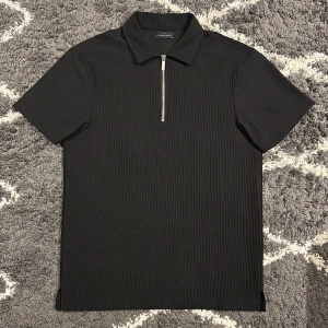 Svart pikétröja - Svart pikétröja från Brand Island i ribbad struktur med snygg silverfärgad dragkedja framtill. Jag är 180cm,67kg och den passar mig bra.