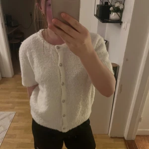 Vit kofta från Gina Tricot - Supermjuk vit kofta från Gina Tricot med kort ärm och pärlknappar framtill. Koftan har en fluffig, stickad känsla och är perfekt att slänga på sig över ett linne eller t-shirt. Enkel och stilren design som passar till allt.