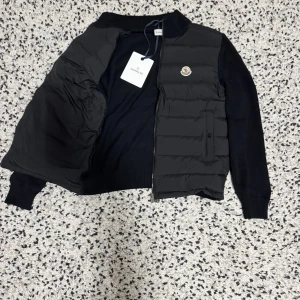 Svart pufferjacka från Moncler - Svart pufferjacka från Moncler med stickade ärmar och quiltad front. Jackan har dragkedja, ribbade muddar och Moncler-logga på bröstet. Perfekt för dig som vill ha en stilren och sportig look med exklusiv känsla.