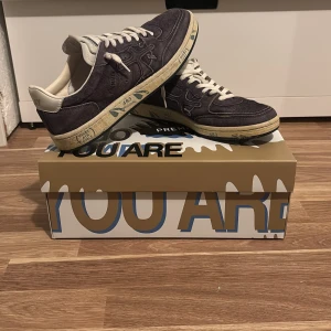 Premiata mörkgråa sneakers mocka - Säljer ett par snygga Premiata sneakers i mörkgråa mocka med beige detaljer och snörning. Skorna har en rund tå och platt sula med coola grafiska tryck på sidan. Perfekt för dig som gillar unik design och vill sticka ut med dina skor. Storlek 41 passar 42. Pris kan diskuteras vid snabb affär.