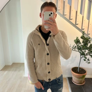 Beige stickad kofta med knappar - Säljer en beige stickad kofta med snygga svarta knappar och ribbad struktur. Koftan har huva, två fickor framtill och en liten logotyp på bröstet. Perfekt för dig som gillar en chill och stilren look.