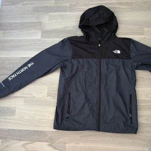 Windbreaker - The North Face - Snygg vindjacka från The North Face i svart och mörkgrått med huva och dragkedja framtill. Jackan har två sidofickor med dragkedja och logga på bröstet och ärmen. Perfekt för blåsiga dagar och outdoor-aktiviteter. Storlek S.