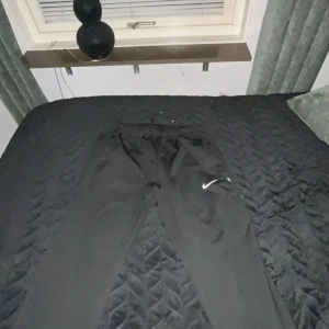 Svarta träningsbyxor från Nike - Svarta träningsbyxor från Nike med elastisk midja och snörning. Byxorna har en vit Nike-logga på ena benet och är tillverkade i ett lätt syntetmaterial. Perfekta för träning eller sportiga outfits.