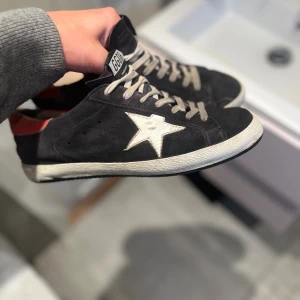 Svarta Golden Goose sneakers - Äkta Golden Goose sneakers i svart mocka med vit stjärndetalj på sidan och röd häl. Skorna har vita snören som är lite smutsiga men kan tvättas om så önskas, vit sula och svart yttersula. Klassisk låg modell. Sulan är i gott skick med många år kvar i sig. Ända som drar ner skicket på skorna är att tyget vid hälen som tyvärr spruckit lite men det är inget som synns vid användning. Använts ca 1 gång i veckan och tagits väl hand om! Hör av dig vid några funderingar! Pris är diskuterbart.