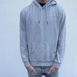 Ljusgrå hoodie med huva och ficka - En stilren ljusgrå hoodie med huva och snörning. Tröjan har en klassisk känguruficka framtill och ribbade muddar vid ärmslut och nederkant. Perfekt för en chill och avslappnad look. Materialet känns mjukt och bekvämt mot huden.