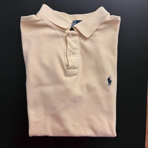 Gul pikétröja från Ralph Lauren - Klassisk gul pikétröja från Ralph Lauren med blå broderad logga på bröstet. Tröjan har krage, knäppning med två knappar och korta ärmar. Tillverkad i mjuk bomull som känns skön mot huden. Perfekt för en avslappnad och stilren look.