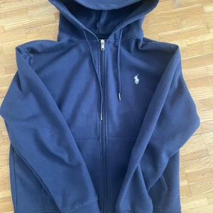 Mörkblå hoodie från Polo Ralph Lauren - Mörkblå hoodie från Polo Ralph Lauren med dragkedja framtill och klassisk huva med snörning. Tröjan har det ikoniska broderade logot på bröstet och är tillverkad i mjuk bomull. Perfekt för en chill och stilren look. Storlek S (170-180 ish), har ingen bild med den på pga att den är liten och har bara legat i garderoben. 