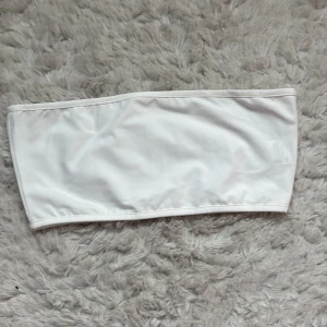Vit bandeautopp från SHEIN - Vit bandeautopp från SHEIN i enkel och stilren design. Tillverkad i stretchigt syntetmaterial som sitter skönt och passar perfekt till nästan alla underdelar. Modellen är axelbandslös och har en clean look utan detaljer. Aldrig använt då den är för stor, nypris 47kr.