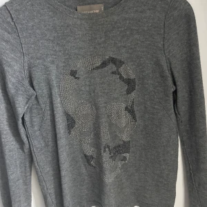 Grå tröja med döskalle från Zadig & Voltaire - Grå långärmad tröja från Zadig & Voltaire med en glittrig döskalle framtill. Tröjan är i kashmir men har ett litet hål på ryggen som behövs sy