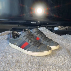 Gucci ace - Gucci sneakers i mörkgrått med klassiskt GG-monogram, röda och blå ränder på sidorna och svarta skosnören. Ovandel i canvas med detaljer i skinn och vit sula. Snygg streetstil och ikonisk design, perfekt för dig som gillar lyxiga sneakers.