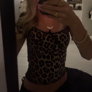 Leopardmönstrad croppad topp - Säljer en croppad topp med leopardmönster och korta ärmar. Toppen har svarta spetsdetaljer längs urringningen och en liten rosett framtill. Sitter tight och har en snygg passform som framhäver midjan. Perfekt för dig som vill sticka ut med ett djurmönstrat plagg.