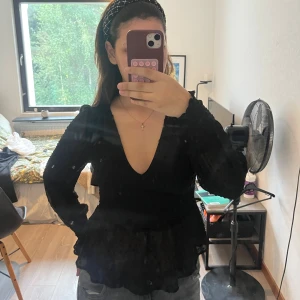 Svart blus med prickig mesh och v-ringning - Säljer en svart blus med transparenta, prickiga meshärmar och djup v-ringning. Blusen har smockade muddar och volang nertill, vilket ger en snygg siluett. Perfekt för dig som gillar trendiga detaljer och en feminin look.