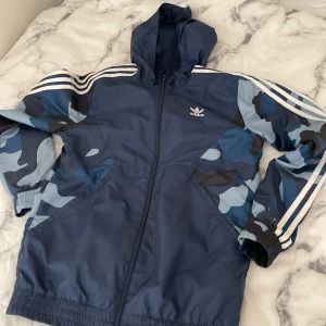 Blå camo vindjacka från Adidas - Snygg blå vindjacka från Adidas med huva och dragkedja. Ärmarna har blått camo-mönster och klassiska vita ränder längs axlarna. Jackan har elastiska muddar och två fickor framtill. Perfekt för dig som gillar sportig streetstyle.